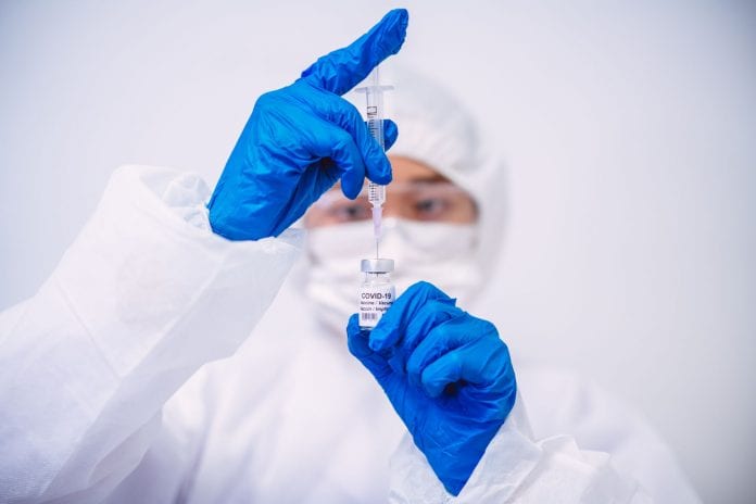 Doctor in protective gloves & workwear filling injection syringe with COVID-19 vaccine